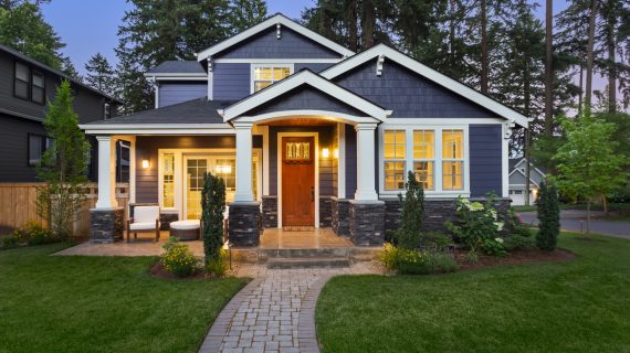 facade of home with manicured lawn, landscaping, and backdrop of trees and dark blue sky. Glowing interior lights create a welcoming mood.