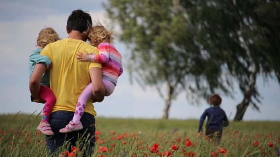 A father carries his two young children through a field of flowers while another child runs ahead.