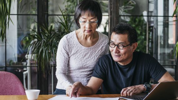 Chinese couple working on home finances with laptop, man wearing glasses and explaining, serious woman watching